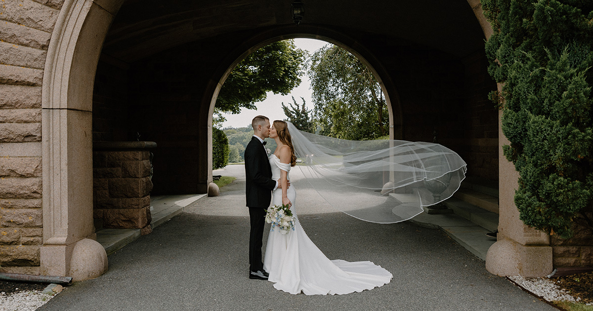 Bride and groom kissing under arch at OceanCliff in Newport, RI, veil blowing in the wind and bride holding flowers